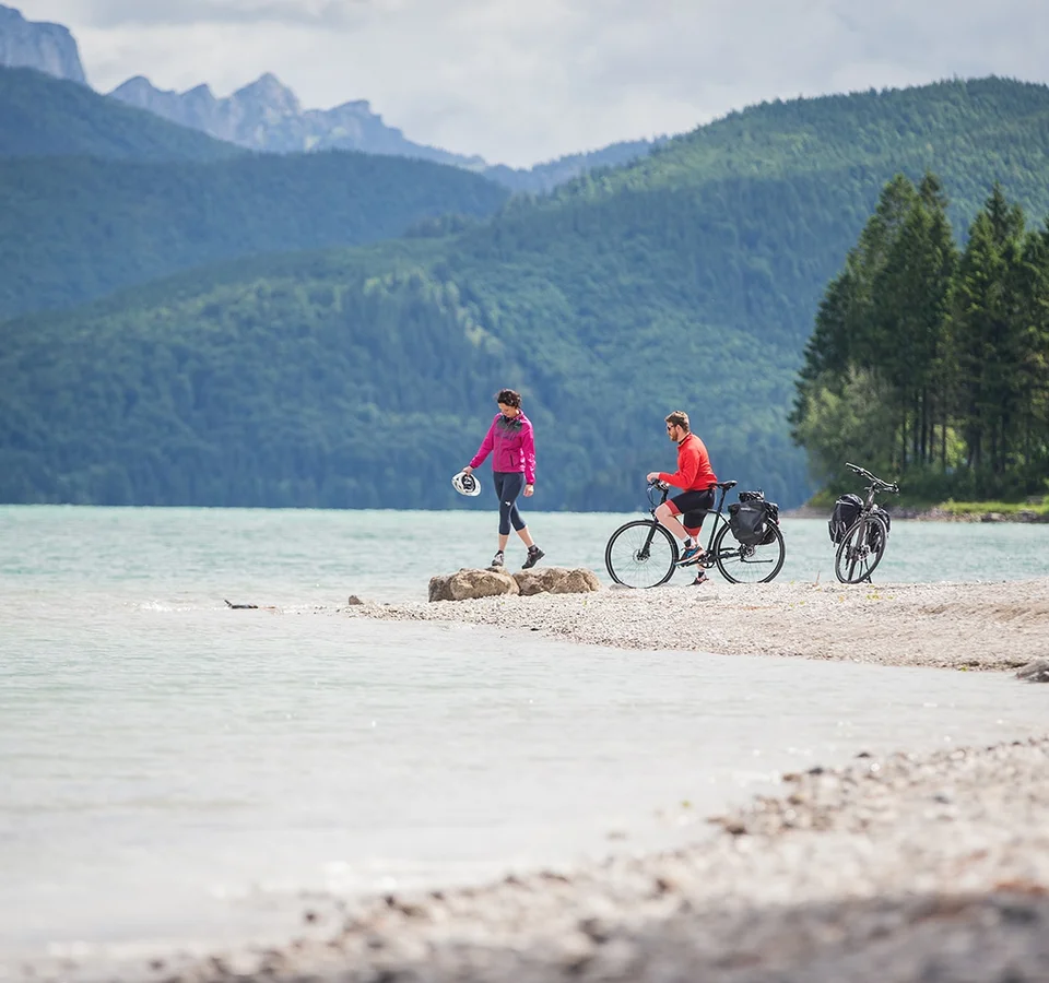 Zwei Radfahrer am Seeufer vor bewaldeten Bergen