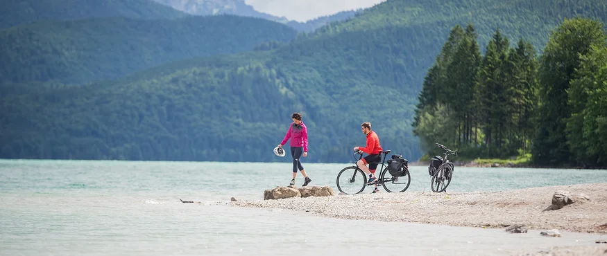 Zwei Radfahrer am Seeufer vor bewaldeten Bergen