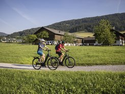 Two cyclists ride on a path through green fields with houses and mountains