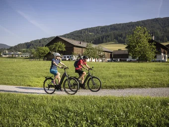 Radstadt © Velontour Two cyclists ride on a path through green fields with houses and mountains