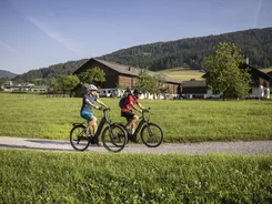 Twee fietsers rijden op een pad door groene velden met huizen en bergen