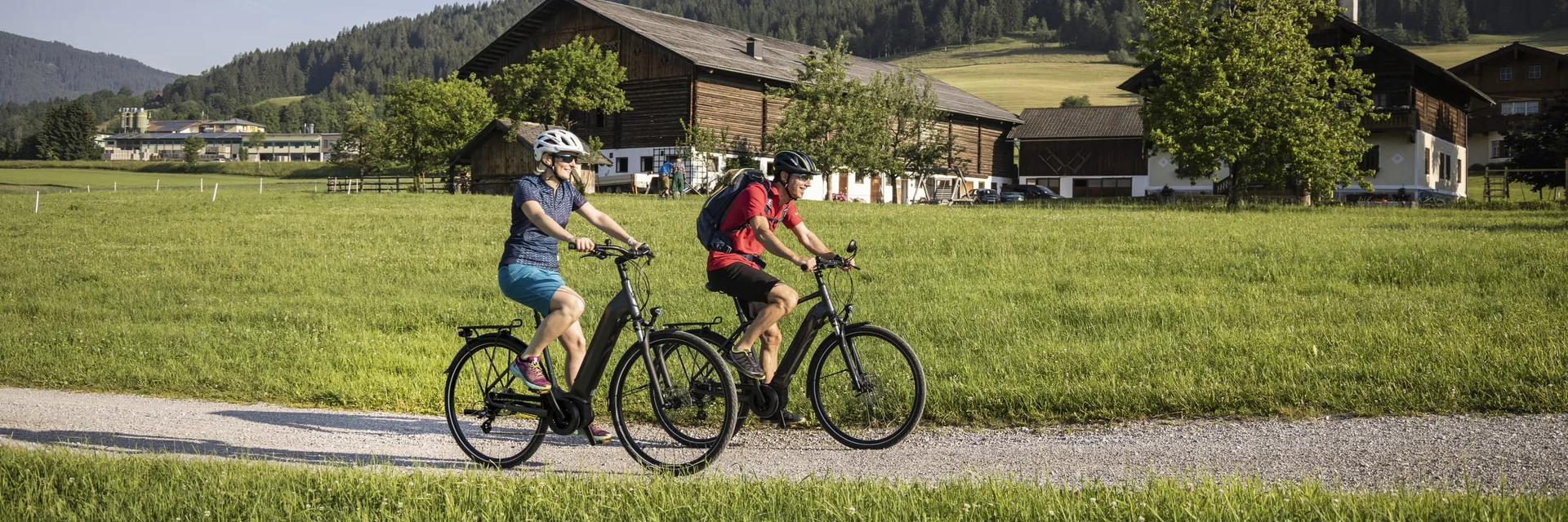 Twee fietsers rijden op een pad door groene velden met huizen en bergen
