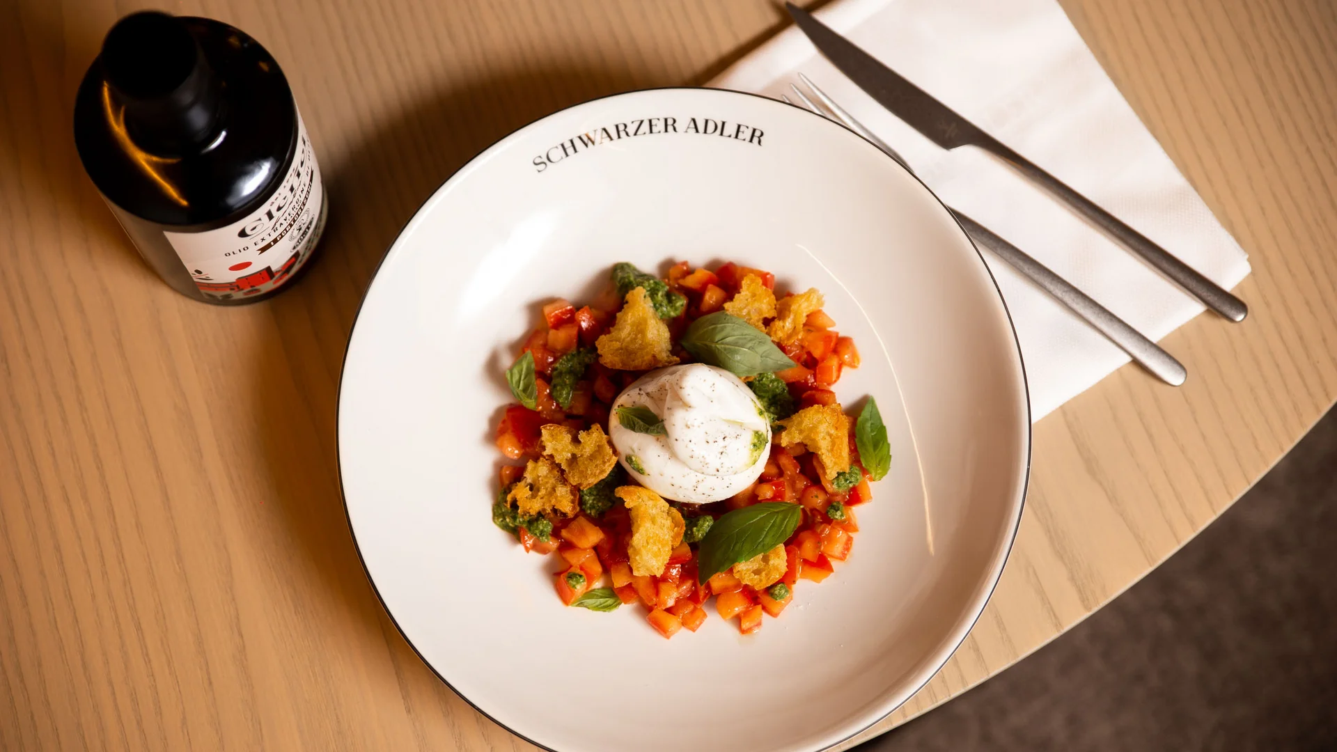 Bord met burrata, blokjes tomaat en basilicum op houten tafel met mes en vork
