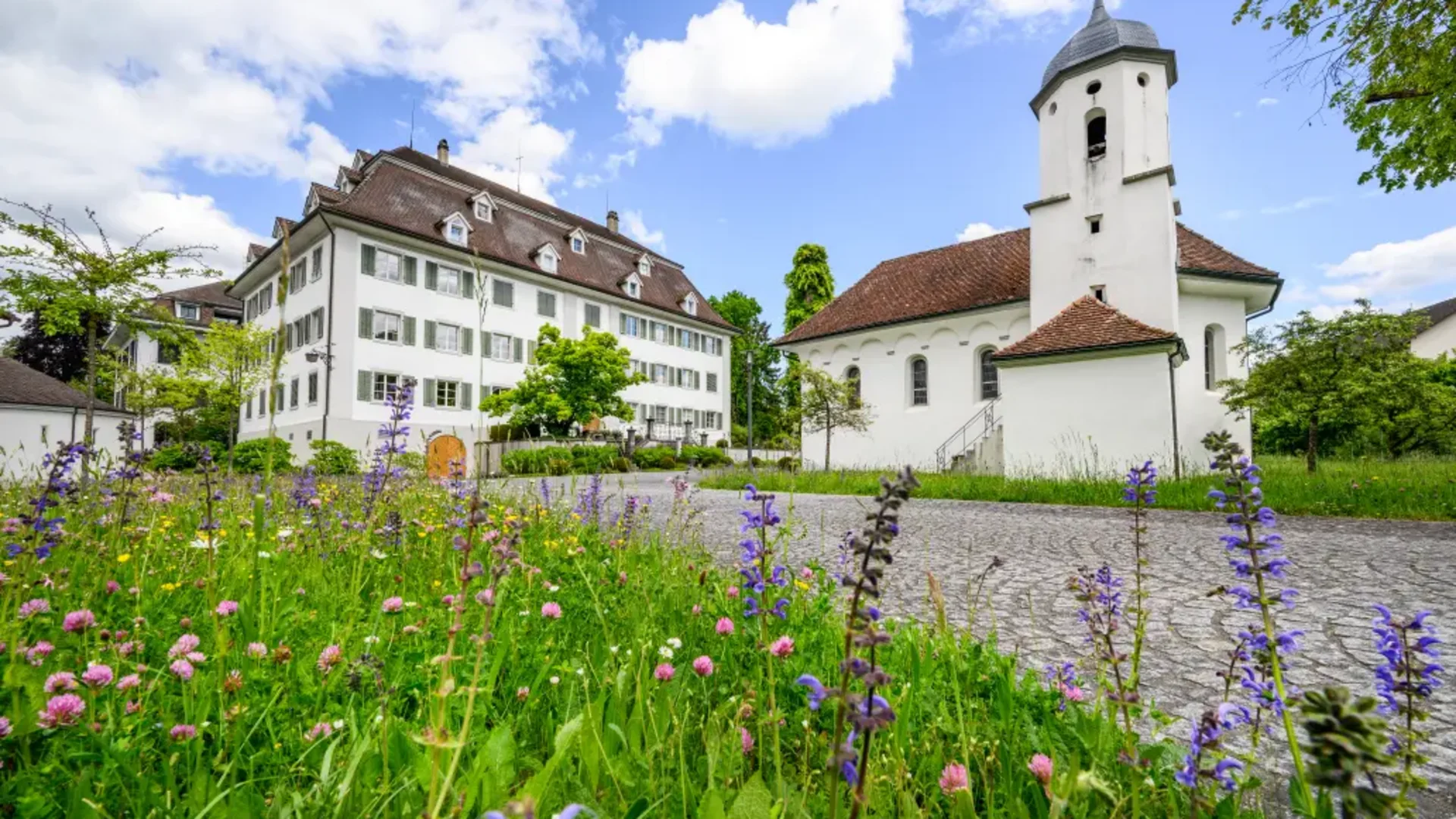 Tour-Detail © Balz Kubli Kirche und altes Gebäude neben Blumenwiese unter blauem Himmel