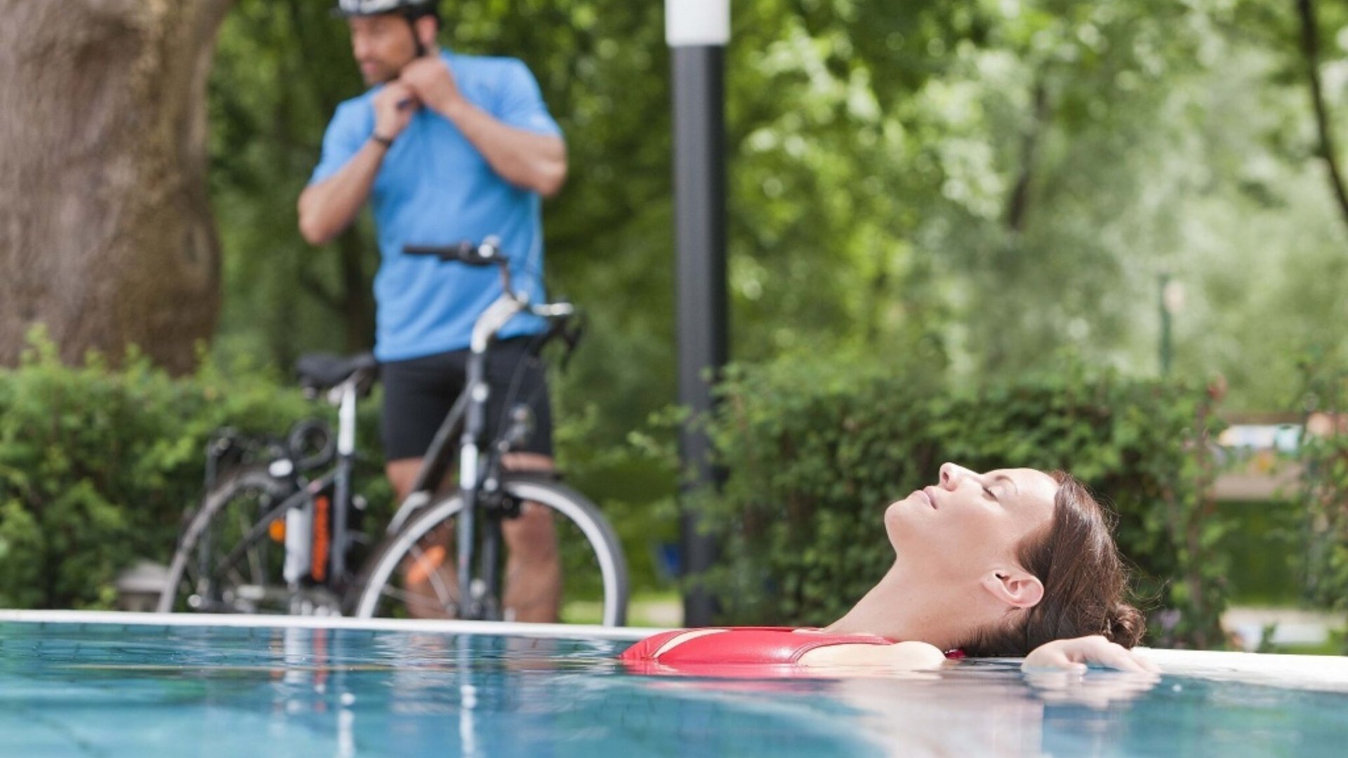 Woman relaxing in pool, man with bicycle in background