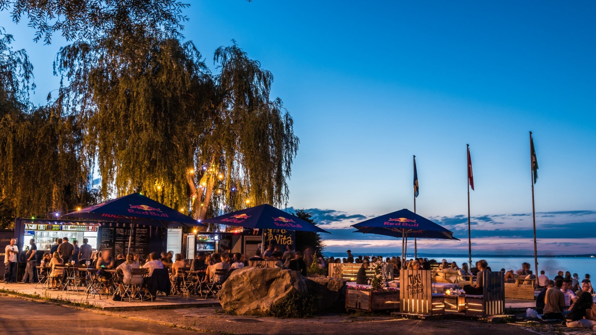 Evening beach bar with guests under Red Bull umbrellas by the lake
