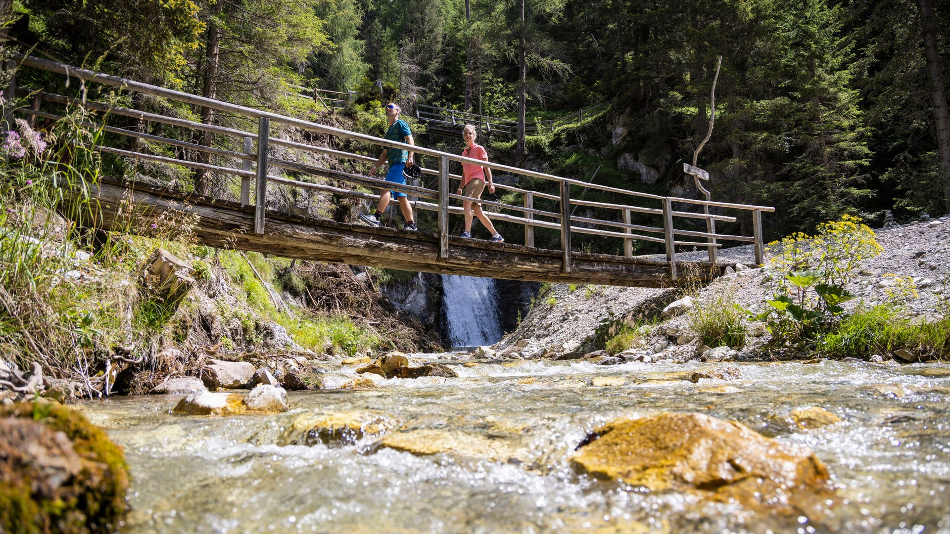 Two hikers crossing a wooden bridge over a clear stream in a forest