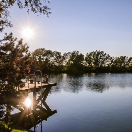 Child sitting on pier by calm lake with sun in background