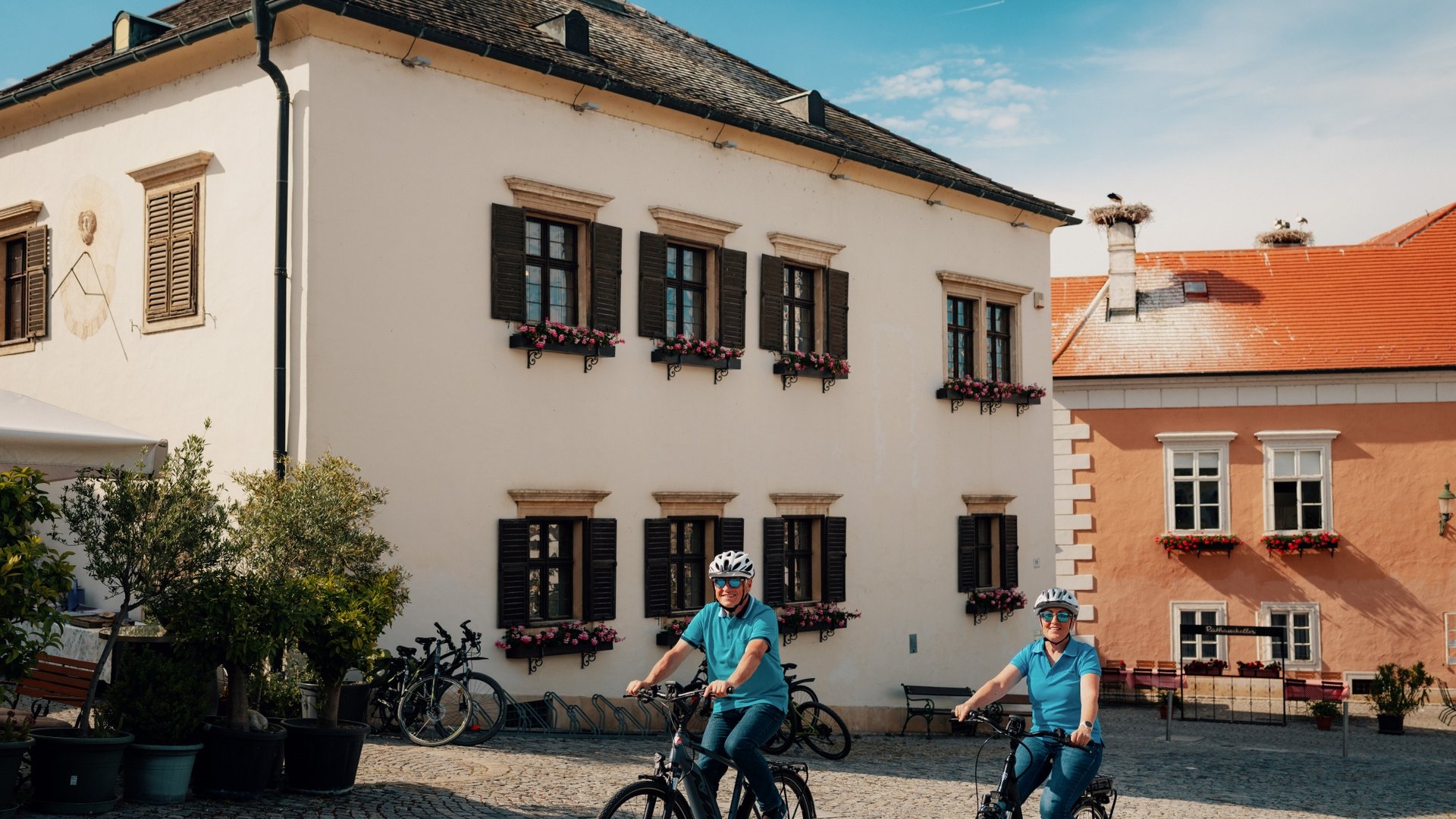 Two people cycling in front of historic buildings on a sunny day