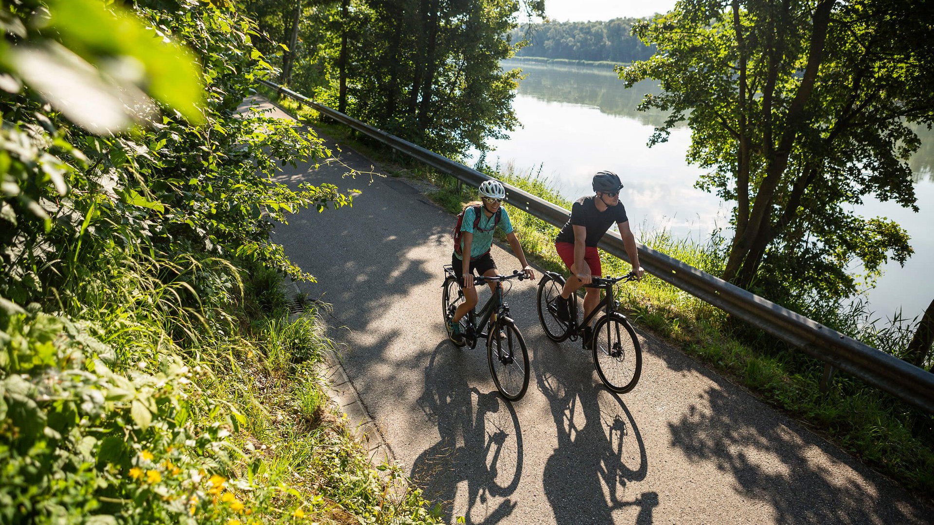 Two cyclists riding on a path beside a river on a sunny day