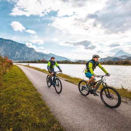 Zwei Radfahrer fahren auf einem Weg entlang eines Flusses mit Bergen im Hintergrund