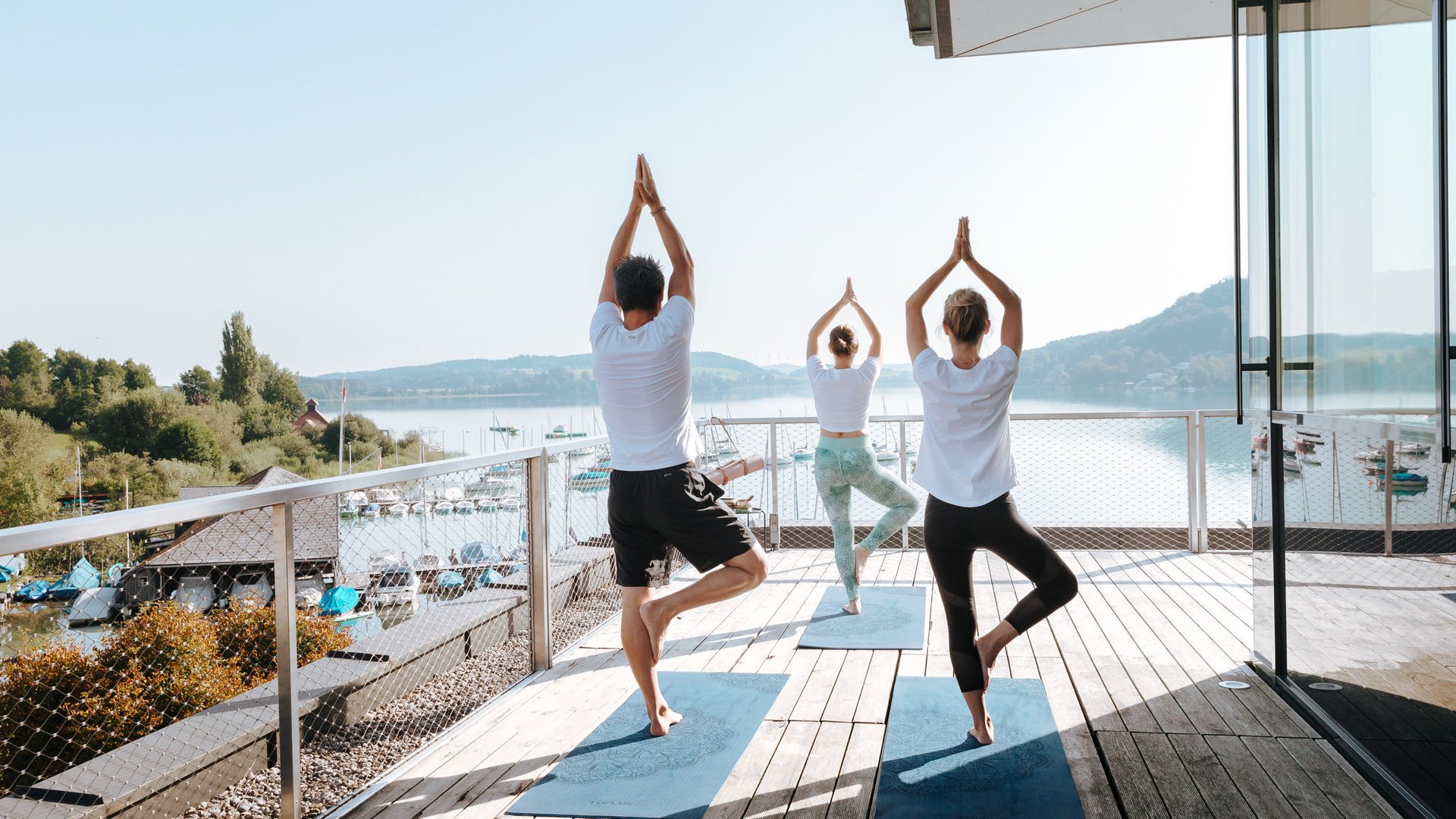 Three people practicing yoga on balcony overlooking lake