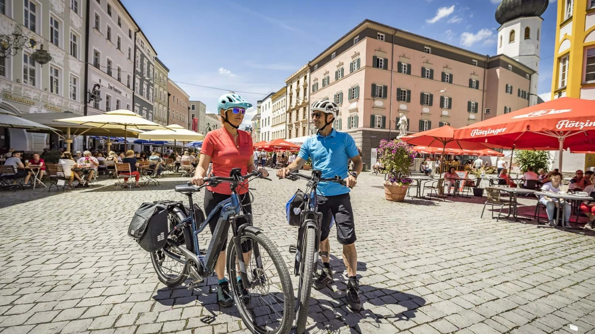 Zwei Radfahrer auf einem gepflasterten Platz mit Cafés und historischen Gebäuden