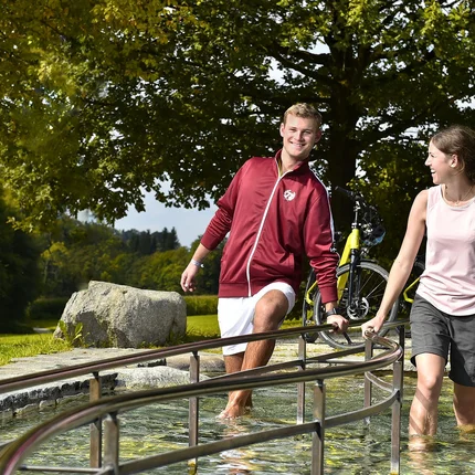 Junge Leute gehen barfuß durch ein Kneippbecken im Park bei Sonnenschein