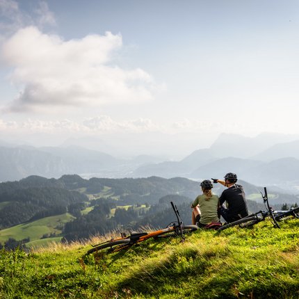 Zwei Radfahrer sitzen auf einem Berg und blicken auf eine bewaldete Alpenlandschaft.