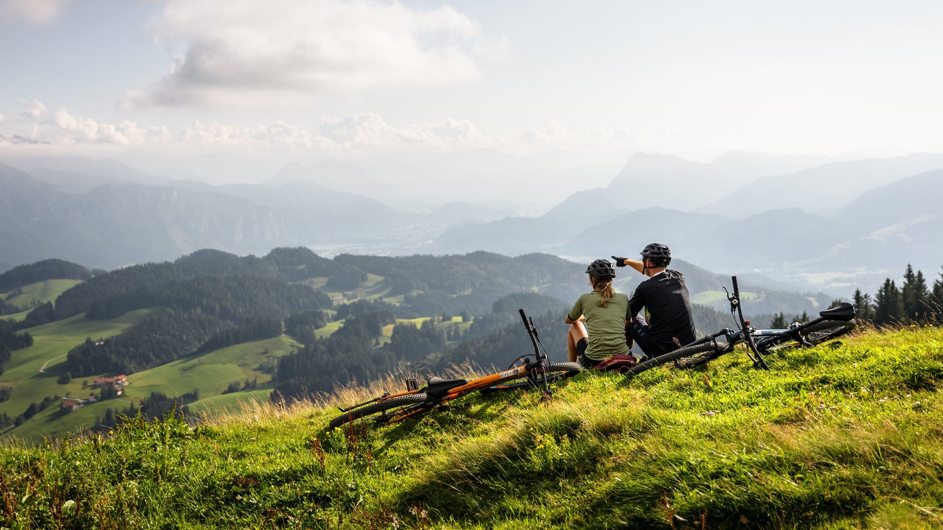 Two cyclists sit on a hill overlooking a forested alpine landscape.