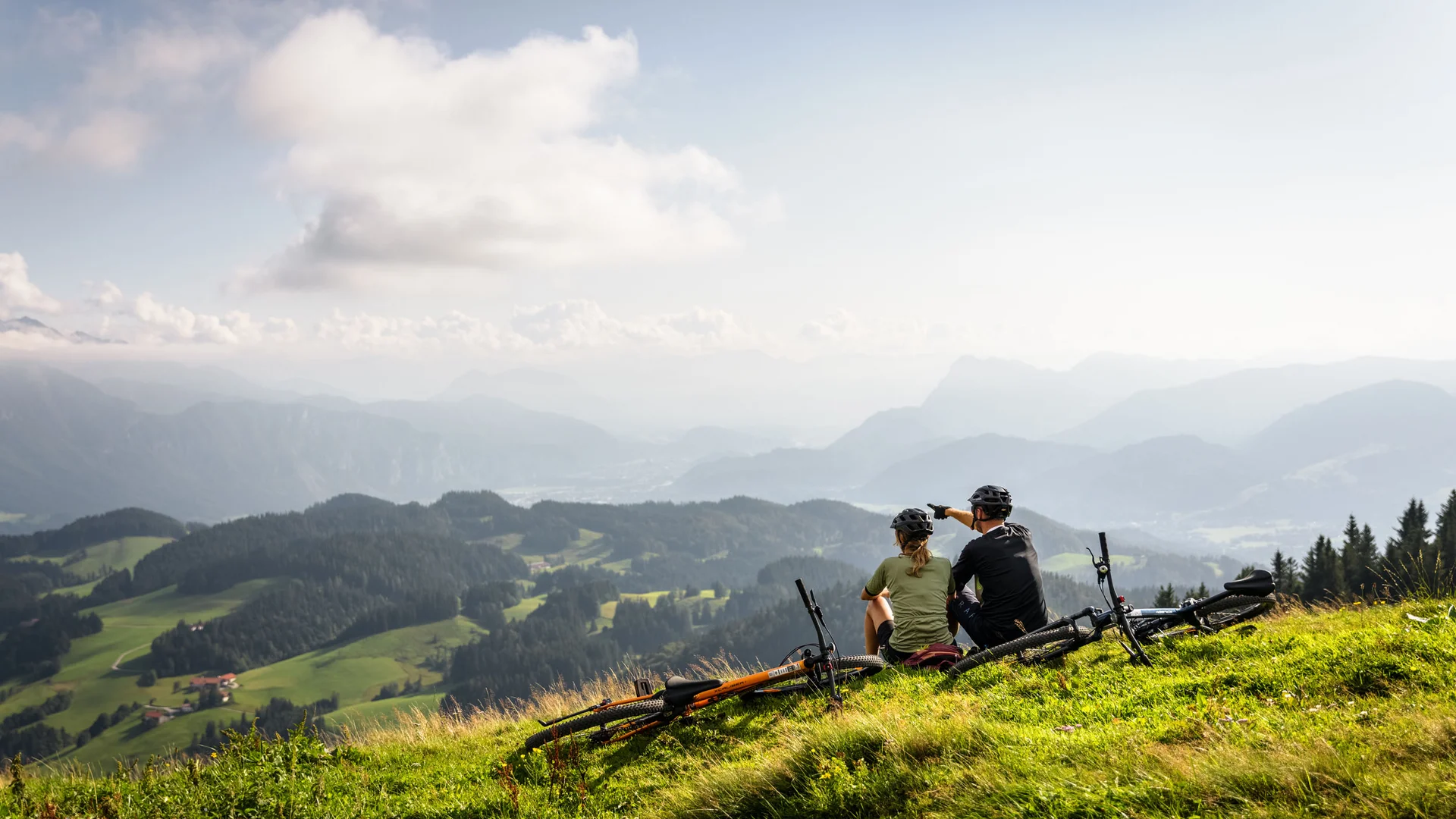 Zwei Radfahrer sitzen auf einem Berg und blicken auf eine bewaldete Alpenlandschaft.