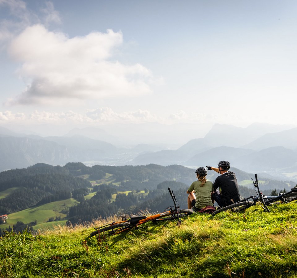 Zwei Radfahrer sitzen auf einem Berg und blicken auf eine bewaldete Alpenlandschaft.