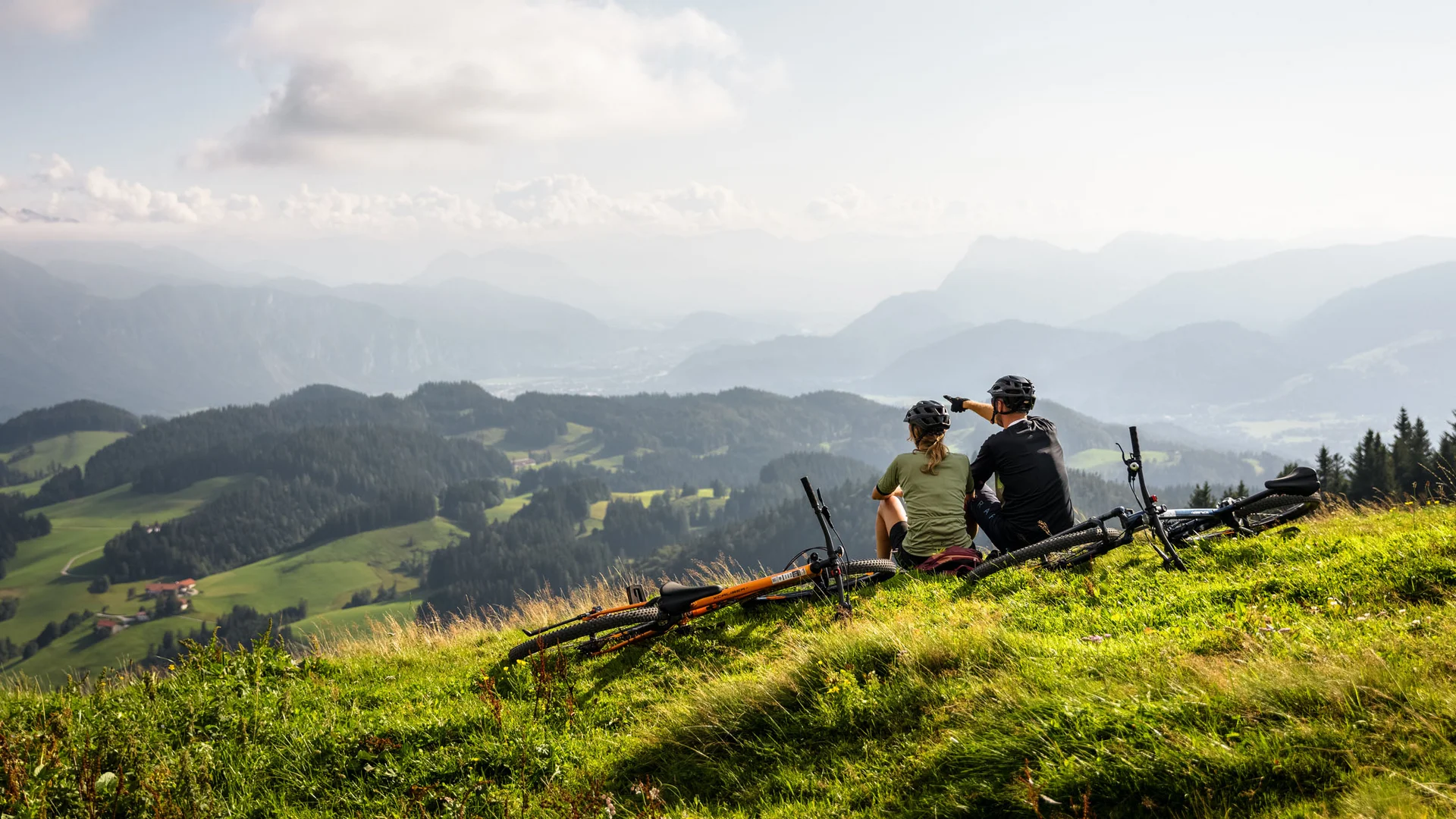 Zwei Radfahrer sitzen auf einem Berg und blicken auf eine bewaldete Alpenlandschaft.