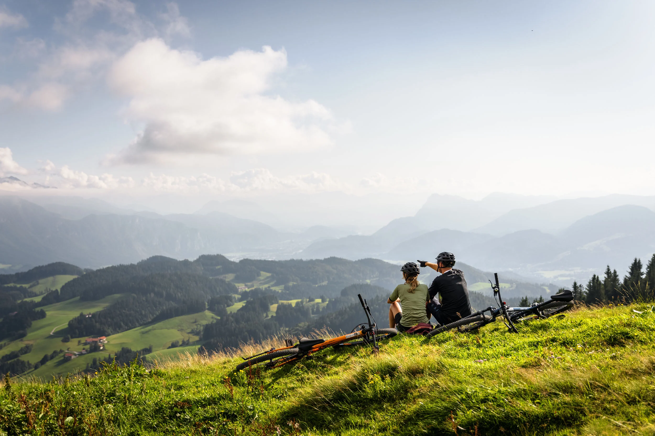 Tirol © Max Dräger Zwei Radfahrer sitzen auf einem Berg und blicken auf eine bewaldete Alpenlandschaft.