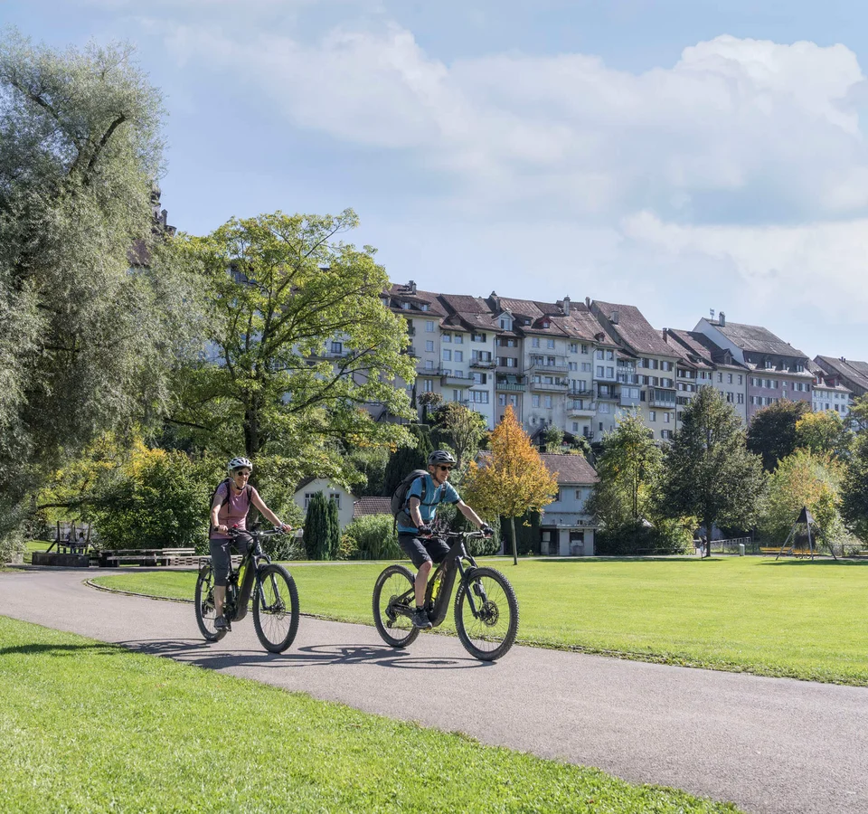 Zwei Radfahrer auf einem Weg in einem Park vor historischen Gebäuden