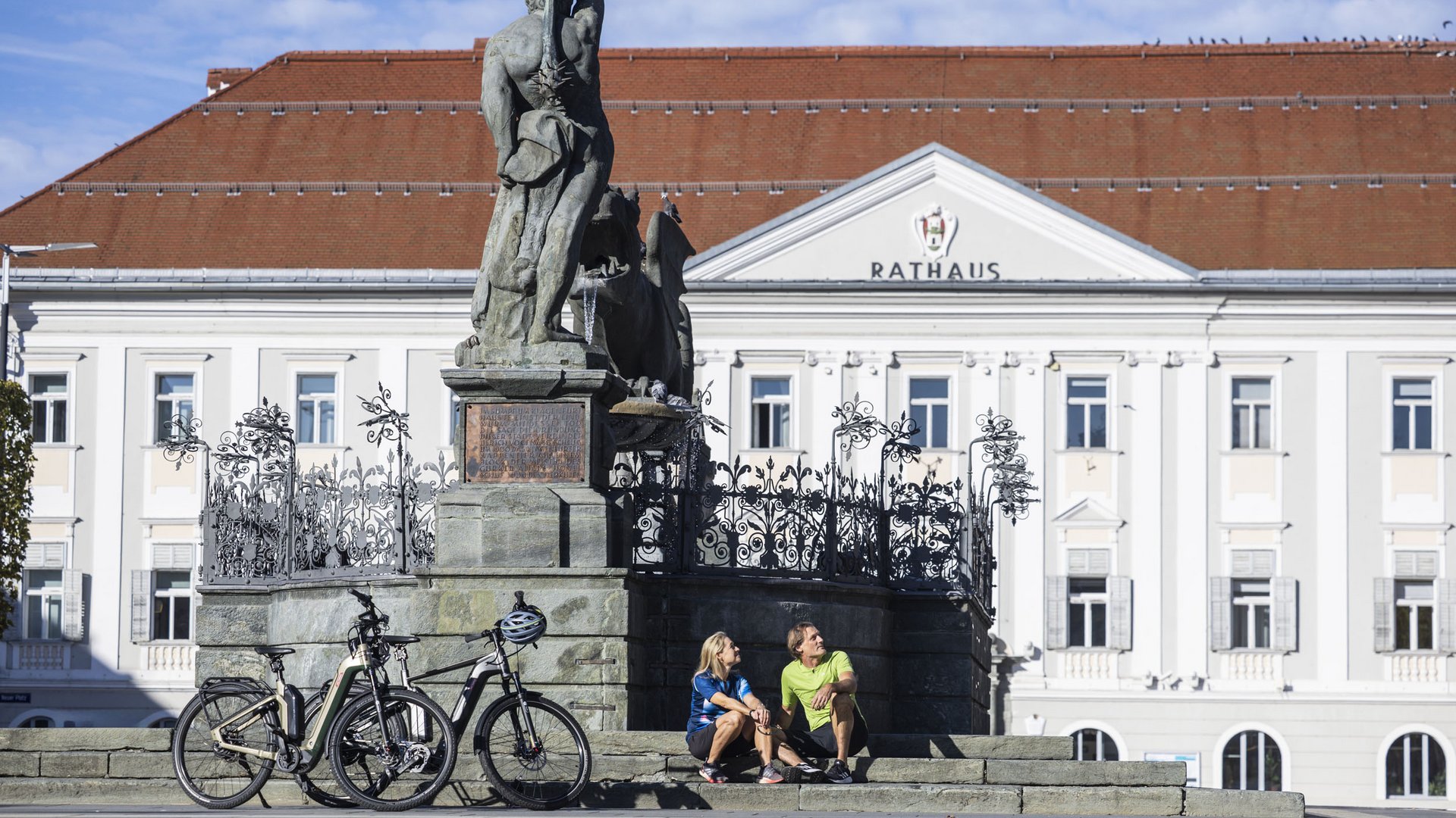 People sitting on steps by statue and bicycles in front of town hall