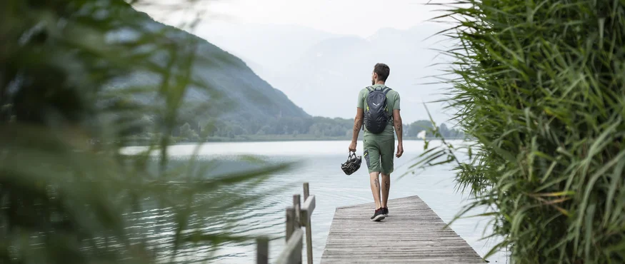 Kulinarisch unterwegs in Tramin © IDM Südtirol - Alex Filz Mann mit Rucksack und Fahrradhelm geht auf Steg am See mit Bergen im Hintergrund