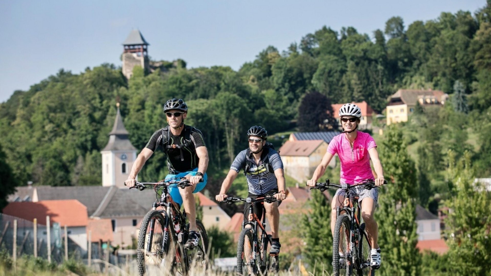 Three cyclists riding on a trail with a village and trees in the background