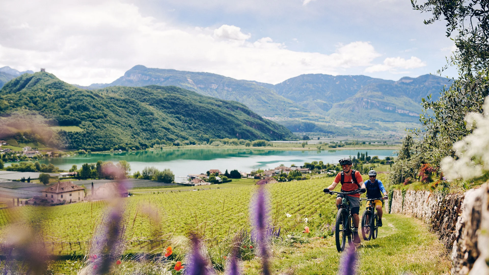 Twee fietsers fietsen door een groen landschap met bergen en meer op de achtergrond