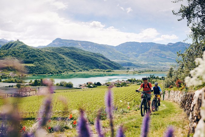 Zwei Radfahrer fahren durch eine grüne Landschaft mit Bergen und See im Hintergrund