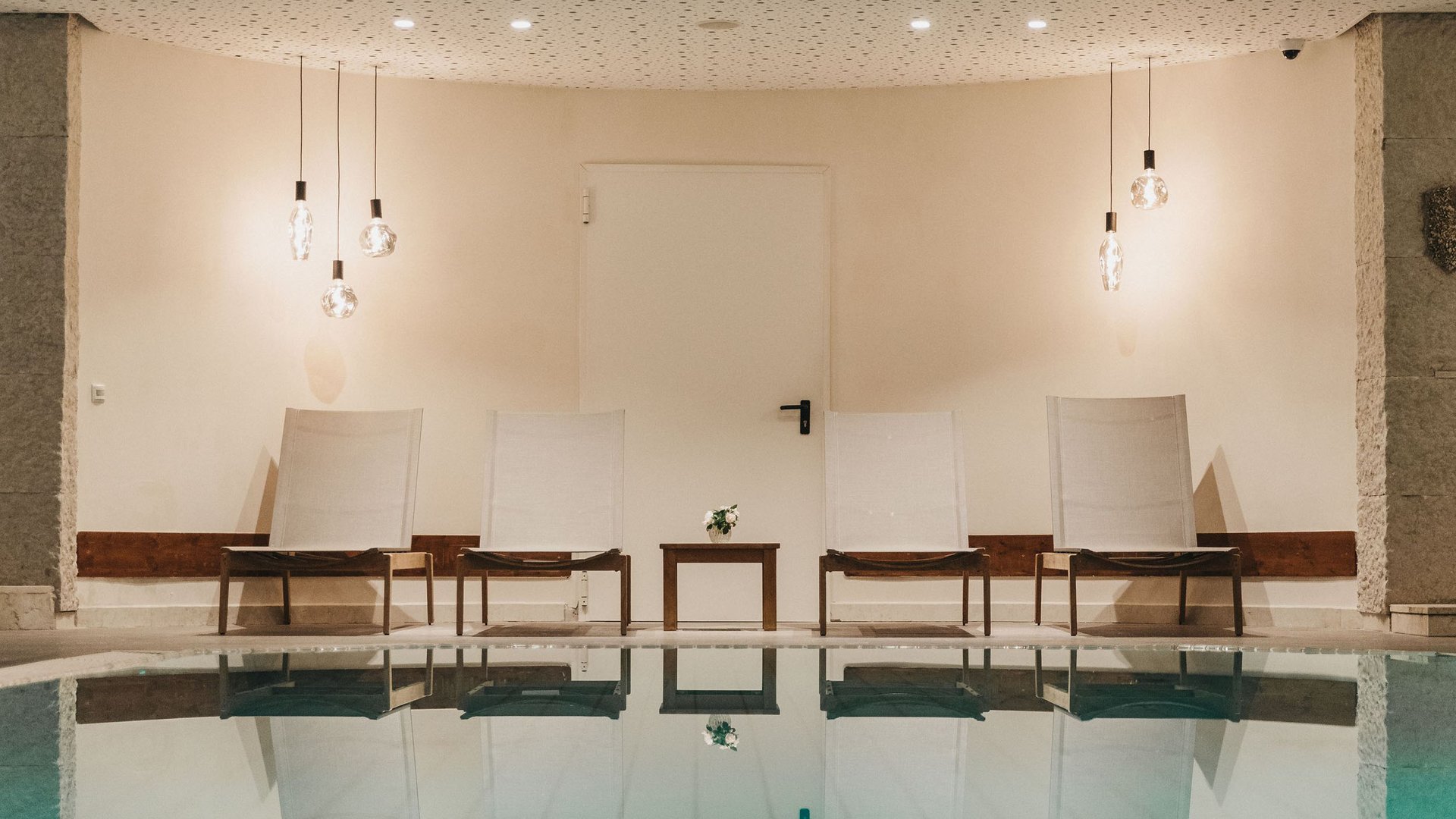 Four white chairs and a small table by a pool with ceiling lights