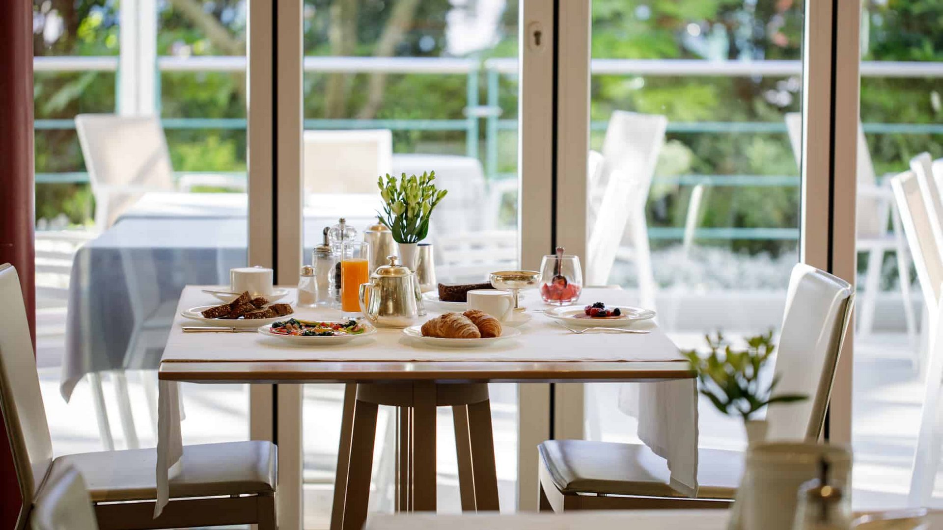 Breakfast table with croissants, juice, and coffee in bright room with glass windows