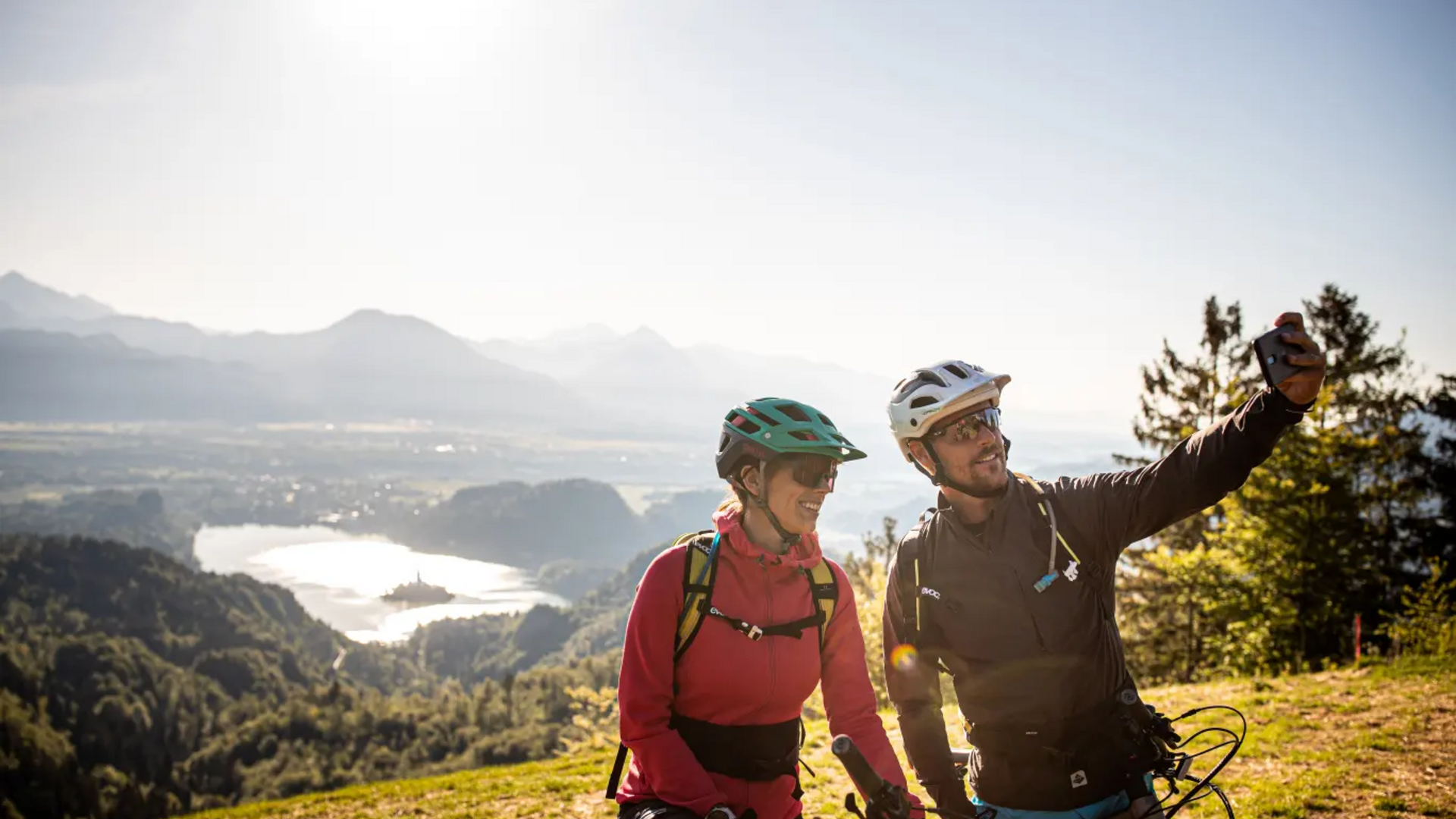 Two cyclists taking a selfie with mountain and lake view in sunlight