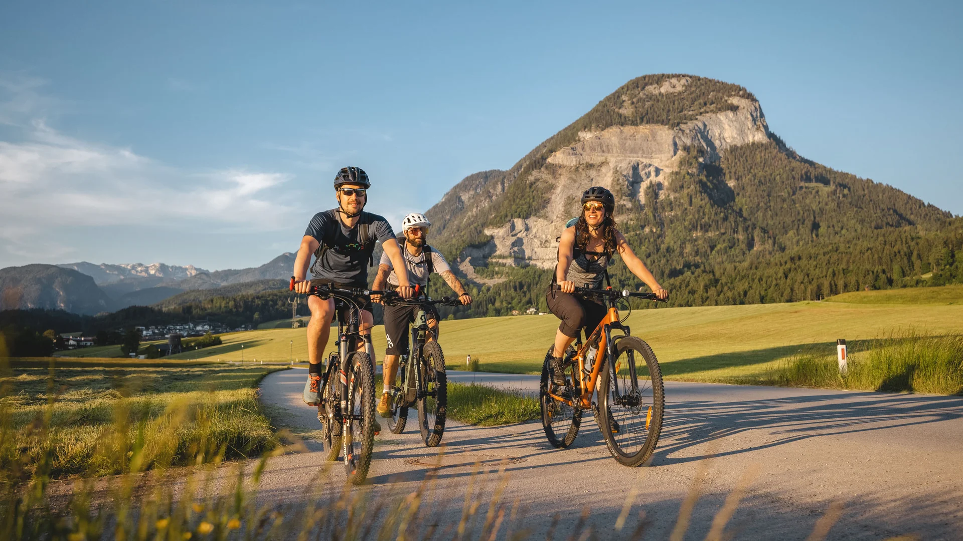 Drei Radfahrer fahren auf Landstraße vor Bergkulisse im Sonnenschein