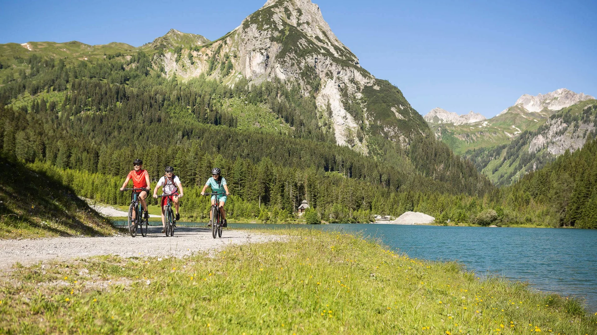 Drei Radfahrer auf einem Weg neben See und Berg im sonnigen Alpengebiet