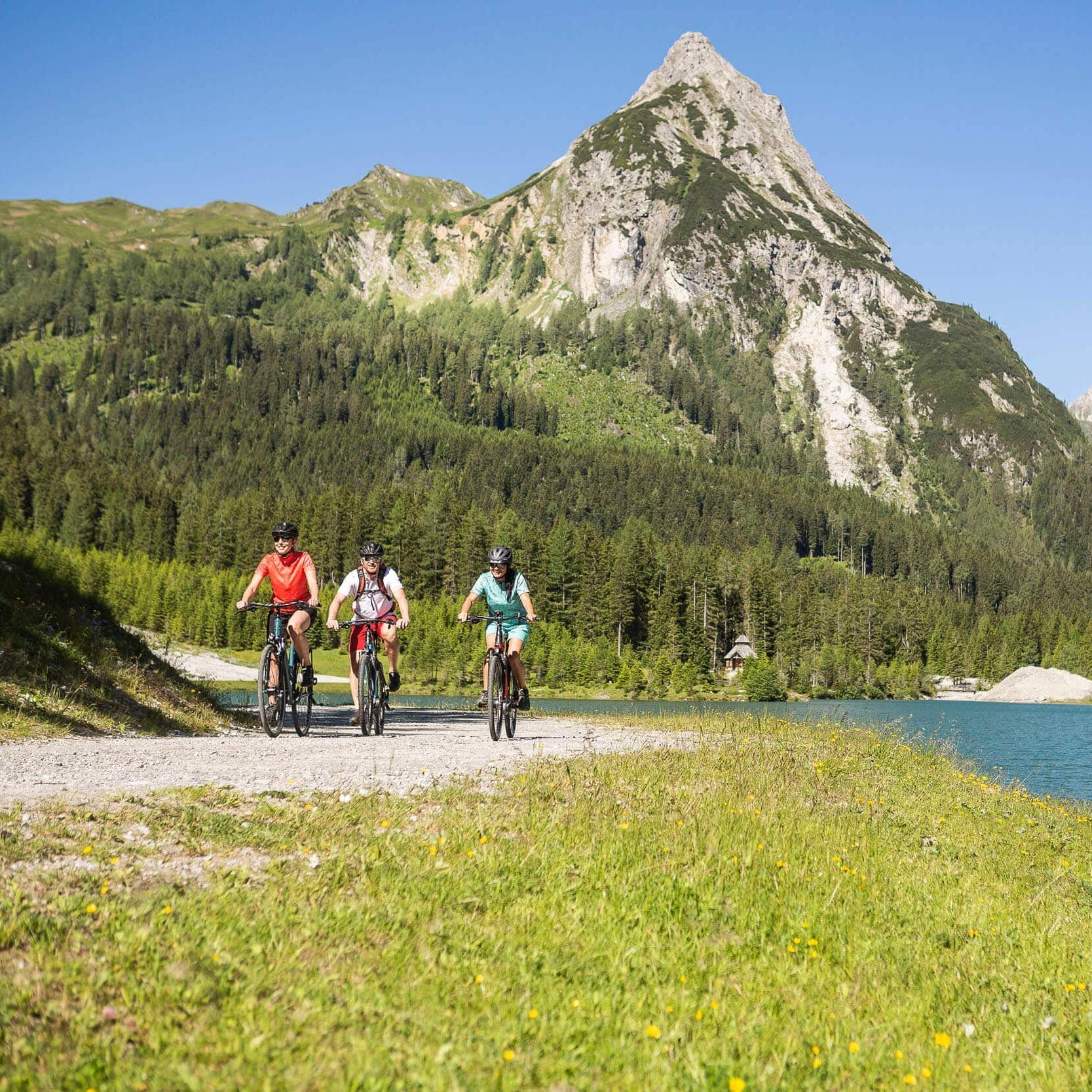 Drei Radfahrer auf einem Weg neben See und Berg im sonnigen Alpengebiet