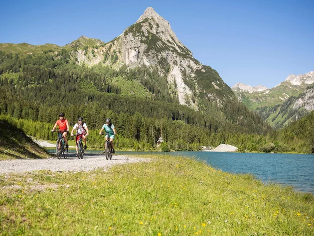 Drei Radfahrer auf einem Weg neben See und Berg im sonnigen Alpengebiet