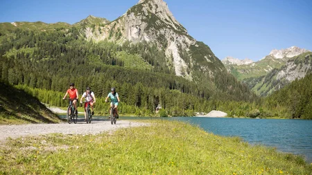 Drei Radfahrer auf einem Weg neben See und Berg im sonnigen Alpengebiet
