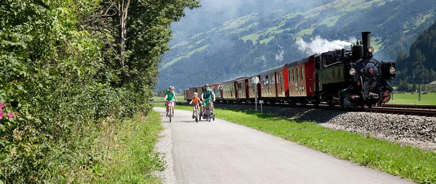 Familie fährt Fahrräder neben historischem Zug in bergiger Landschaft
