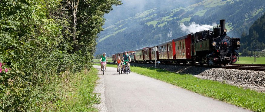 Family cycling beside a historic train in mountainous scenery