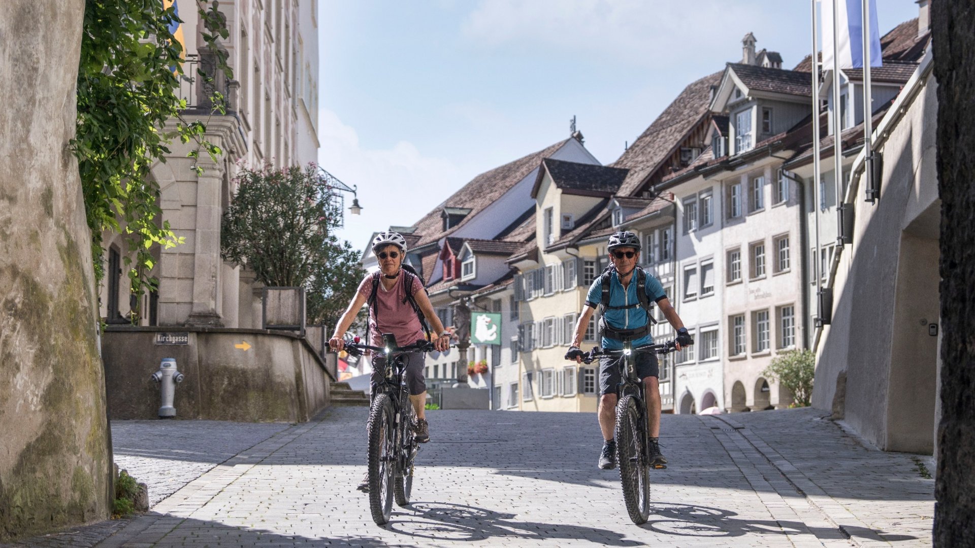 Two adults wearing helmets cycling through an old town street