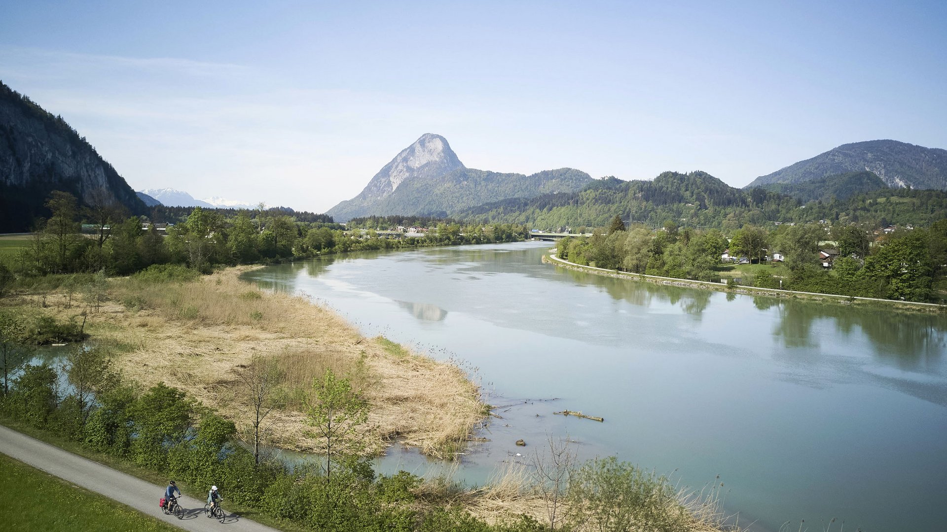 Two cyclists on a path beside a river with mountains in the background