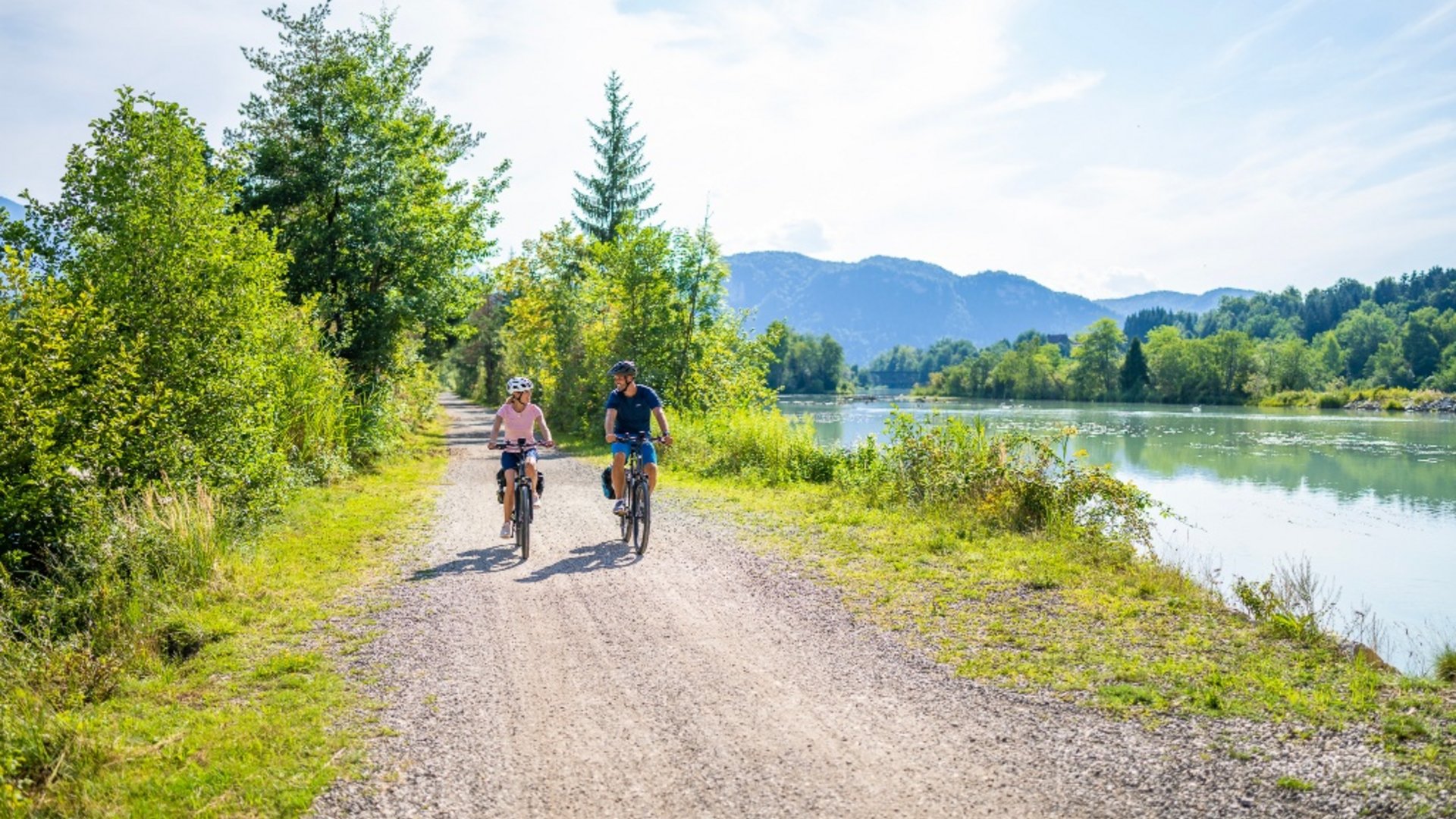Two cyclists riding on a path beside a river in a green landscape