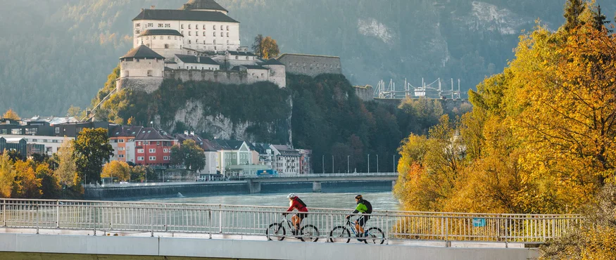 Zwei Radfahrer auf einer Brücke mit Burg und herbstlichen Bäumen im Hintergrund