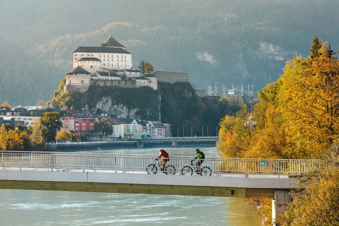 Zwei Radfahrer auf einer Brücke mit Burg und herbstlichen Bäumen im Hintergrund