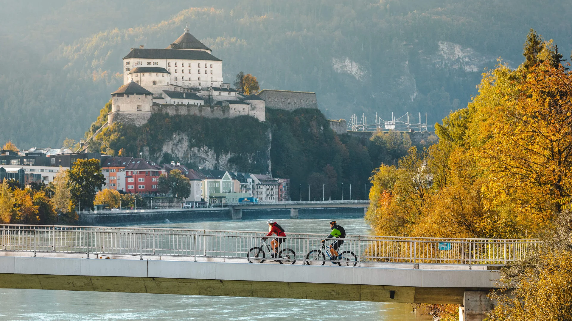 Zwei Radfahrer auf einer Brücke mit Burg und herbstlichen Bäumen im Hintergrund