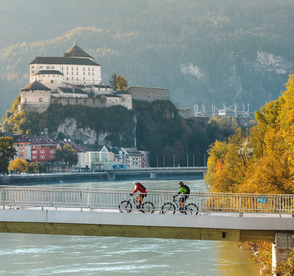 Zwei Radfahrer auf einer Brücke mit Burg und herbstlichen Bäumen im Hintergrund