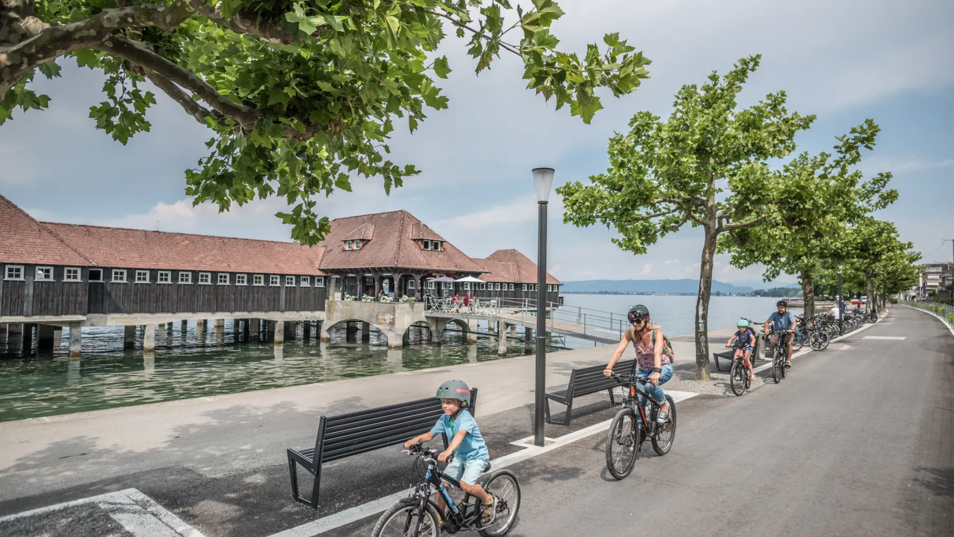 Family cycling on lakeside promenade past historic wooden building over water