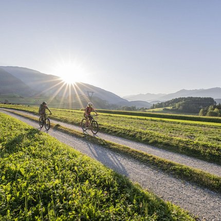 Two cyclists riding on a dirt path with mountains and setting sun in the background