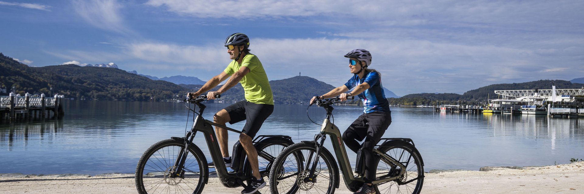 Two people riding e-bikes by a lake with mountains in the background